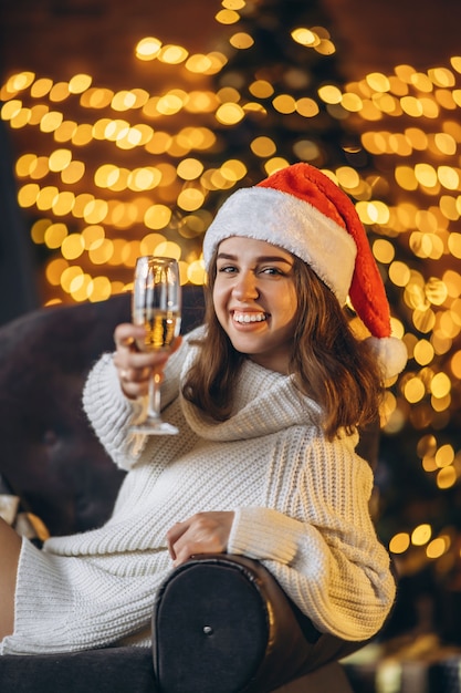 Ładna kobieta w ciepłym swetrze, skarpetkach i świątecznej czapce, siedząca na krześle z szampanem