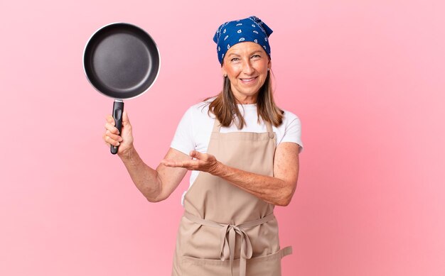 Ładna kobieta w średnim wieku, uśmiechając się radośnie, czując się szczęśliwa, pokazując koncepcję i trzymając patelnię. koncepcja szefa kuchni