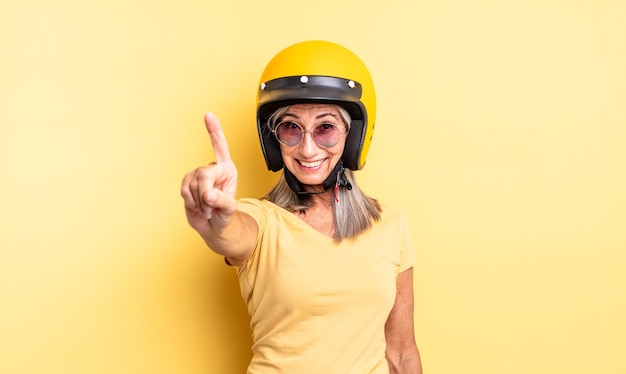 Ładna kobieta w średnim wieku, uśmiechnięta i wyglądająca przyjaźnie, pokazując numer jeden. koncepcja kasku motocyklowego