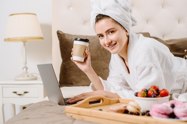 Ładna kobieta w szlafroku i ręczniku na głowie, korzystająca z laptopa na łóżku w domu