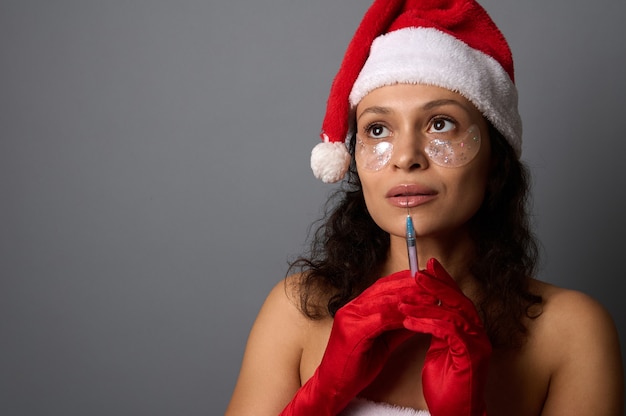 Ładna kobieta z naturalnym makijażem i łatami pod oczami, w Santa hat trzyma strzykawkę z zastrzykiem piękna w pobliżu jej twarzy, patrzy na przestrzeń kopii szarego tła. Kosmetologia wtryskowa, koncepcja spa