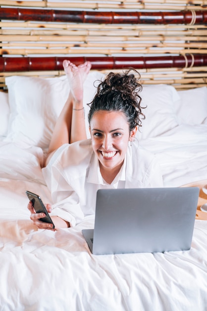 Ładna kobieta z telefonem i laptopem w łóżku