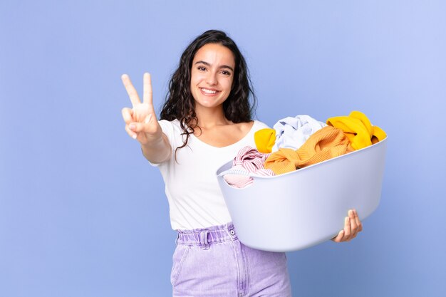 Ładna latynoska kobieta uśmiechnięta i wyglądająca przyjaźnie, pokazująca numer dwa i trzymająca kosz na pranie