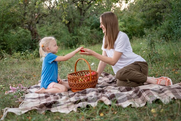 Ładna mała dziewczynka daje jabłku jej matkę lub siostrę na polanie w lesie Letni piknik rodzinny mama i dziewczyna w parku