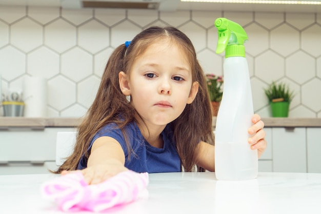 Ładna mała dziewczynka z opryskiwaczem detergentu i stołem do wycierania ragami gospodarstwa domowego Dziecko sprząta w domu sprzątanie kuchni