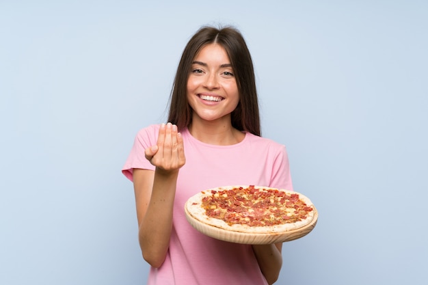 Ładna Młoda Dziewczyna Trzyma Pizzę Nad Odosobnioną Błękit ścianą Zaprasza Przychodzić Z Ręką. Cieszę Się, że Przyszedłeś