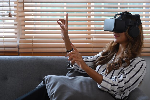 Ładna młoda kobieta ogląda wideo lub gra w gry wideo na zestawie wirtualnej rzeczywistości i dotyka w powietrzu