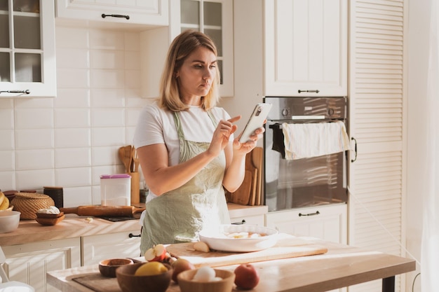 Ładna młoda kobieta przygotowuje szarlotkę w domu w przytulnej kuchni i robi zdjęcia telefonem