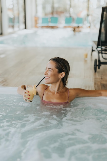 Ładna młoda kobieta relaksująca się w krytym basenie ze szklanką świeżego soku pomarańczowego