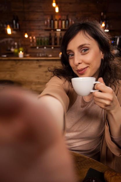 Ładna młoda kobieta robi sef-potrait w pięknej stołówce. Piękna kobieta uśmiechając się do kamery. Piękna kobieta trzyma filiżankę mieszanej kawy.