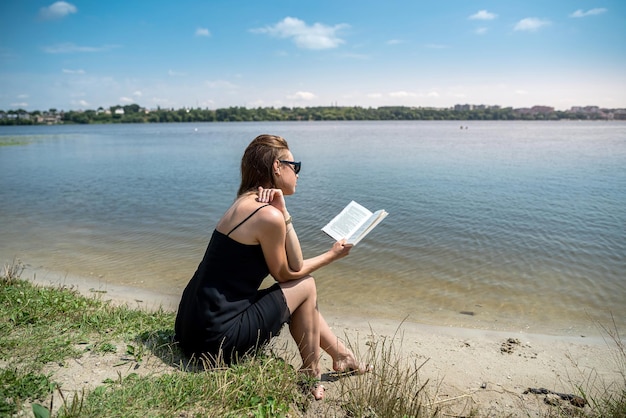 Ładna młoda kobieta w letni dzień czyta książkę w pobliżu jeziora