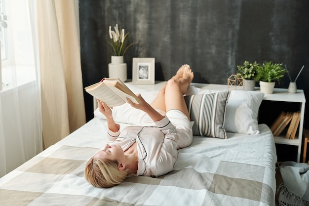 Ładna młoda kobieta w piżamie, relaksując się na łóżku w czasie wolnym i czytając książkę podczas pobytu w domu w weekend
