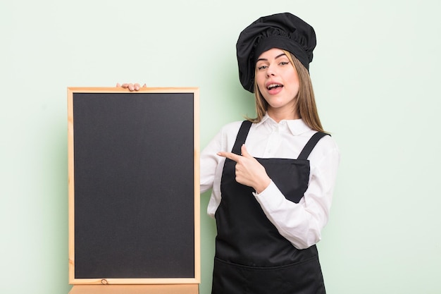 Ładna młoda kobieta wyglądająca na podekscytowaną i zaskoczoną, wskazując na bocznego szefa kuchni z tablicą