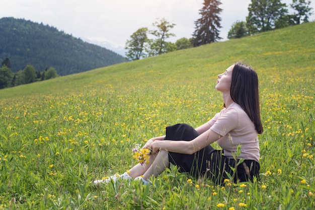 Ładna młoda kobieta z bukietem wildflowers siedzi na gazonie. Góry w tle. Letni słoneczny dzień