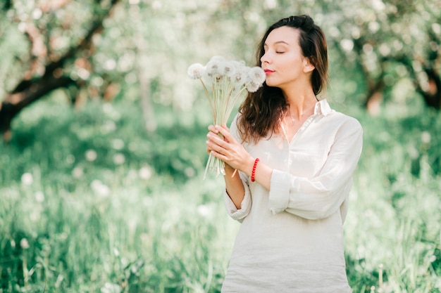 Ładna młoda szczęśliwa brunetki kobieta w białej koszula wącha bukiet dandelions