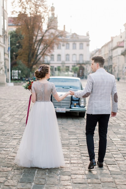 Ładna Panna Młoda I Przystojny Pan Młody Spaceru W Starym Centrum Miasta, Stojący Przed Czarnym Samochodem Retro