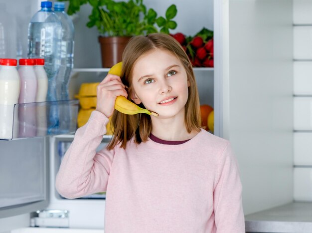 Ładna preteen dziewczyna trzyma banana jako telefon w kuchni w domu i patrzy w kamerę Piękne dziecko płci żeńskiej bawi się owocami