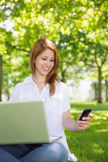 Ładna rudzielec używa jej laptop podczas gdy texting w parku