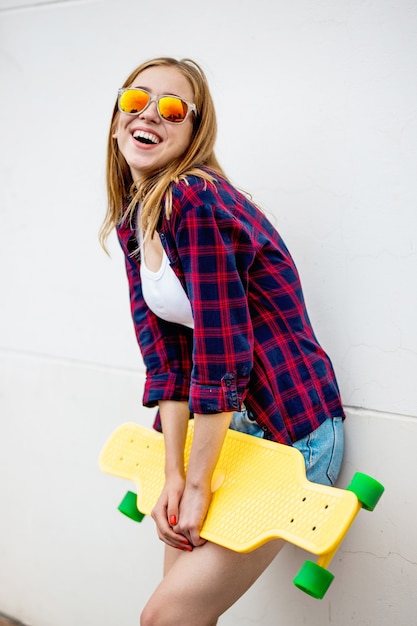 ładna uśmiechnięta blond dziewczyna stoi przed szarą ścianą i trzyma żółty longboard