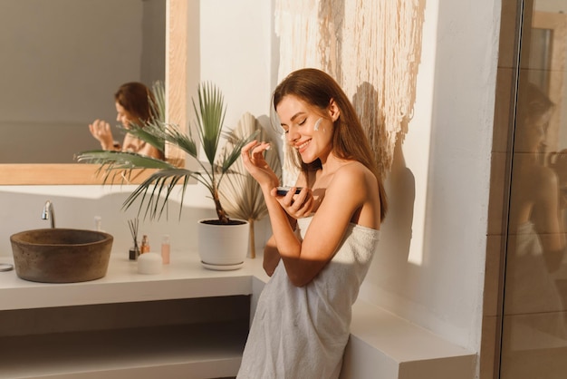 Zdjęcie Ładna uśmiechnięta pani nakłada krem nawilżający na twarz w nowoczesnej łazience