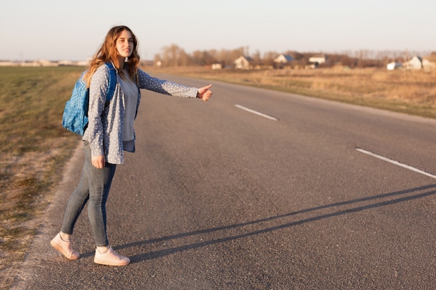 ładna uśmiechnięta podróżniczka stoi na drodze, jeździ autostopem i zatrzymuje samochody, nosi torbę, cieszy się darmowym stylem życia