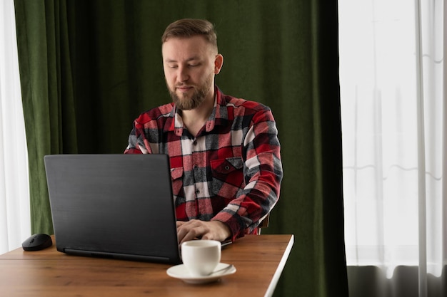 Ładny, brodaty freelancer pracujący w kawiarni z laptopem, poranna praca z filiżanką kawy w przytulnym biurze domowym, praca online.