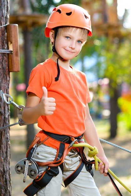 Ładny chłopak pokazuje kciuk ze sprzętem wspinaczkowym w parku rozrywki