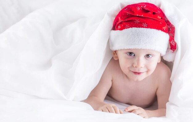 Ładny chłopczyk z santa hat na łóżku pod białą kołdrą.