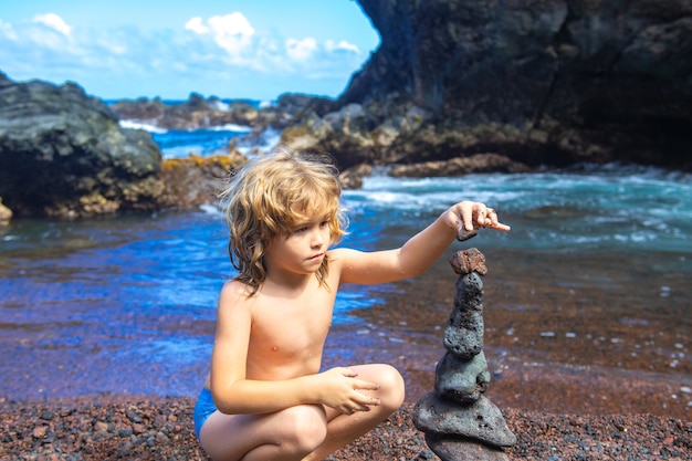 Ładny Chłopiec Dziecko Dokonywanie Stos Kamieni Na Wybrzeżu Morza W Naturze. Kopiec Na Plaży Nad Oceanem, Wieża Z Pięciu Kamyków. Pojęcie Równowagi I Harmonii. Spokój I Duch.