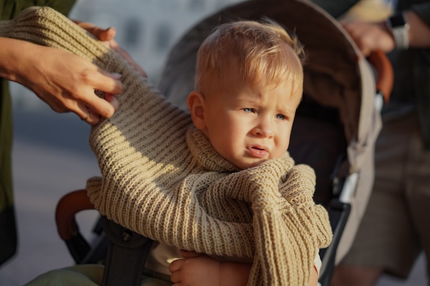 Zdjęcie Ładny kaukaski chłopczyk siedzi w wózku. matka stawia na niego sweter. obraz z selektywnym naciskiem. zdjęcie wysokiej jakości