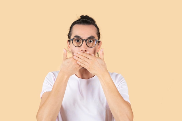 Ładny łaciński mężczyzna z okularami do widzenia zakrywa usta twarz zaskoczenia lub przerażenia beżowego tła