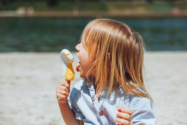 Ładny mały blond chłopiec z długimi włosami, jedzenie lodów na wakacjach na plaży