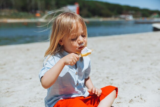 Ładny mały blond chłopiec z długimi włosami, jedzenie lodów na wakacjach na plaży
