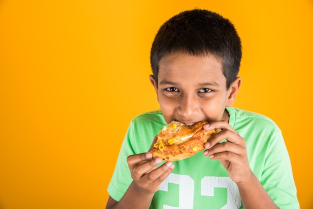 Ładny mały chłopiec indyjski lub azjatycki jedzenie smaczne hamburgery, kanapki lub pizzę w talerzu lub w pudełku. Stojący na białym tle na niebieskim lub żółtym tle.