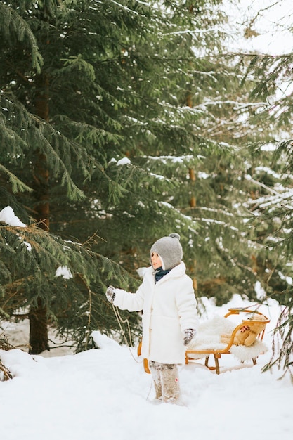Ładny mały chłopiec maluch w futrzanej czapce i filcowych butach siedzi na sankach z misiem zabawka w śniegu zima koncepcja lasu świąt Bożego Narodzenia i nowego roku w stylu retro vintage