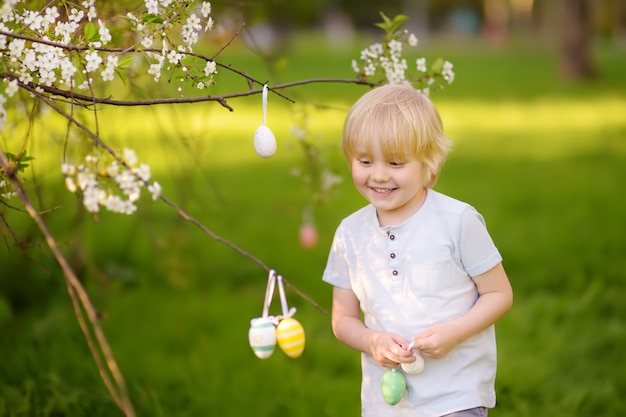 Ładny mały chłopiec poluje na pisanki na gałęzi kwitnących drzew.