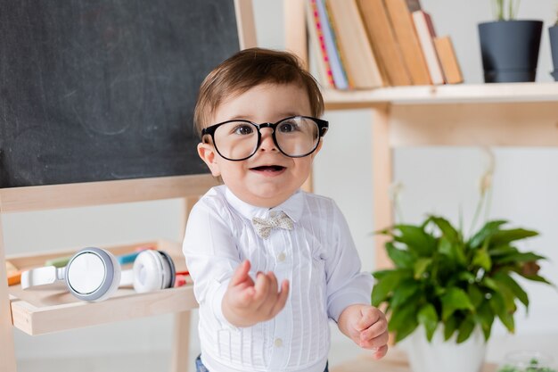 Ładny mały chłopiec z okularami uśmiecha się obok tablicy i książek. edukacja przedszkolna dla dzieci