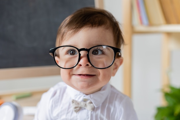 Ładny mały chłopiec z okularami uśmiecha się obok tablicy i książek. edukacja przedszkolna dla dzieci