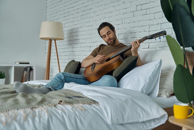 Ładny młody człowiek grający na gitarze akustycznej podczas odpoczynku w domu