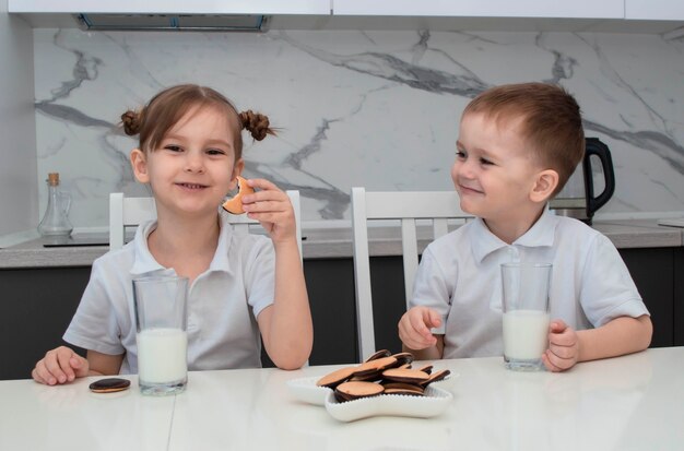 Ładny szczęśliwy mały blond brat i siostra śmieją się i jedzą ciasteczka siedząc przy stole.