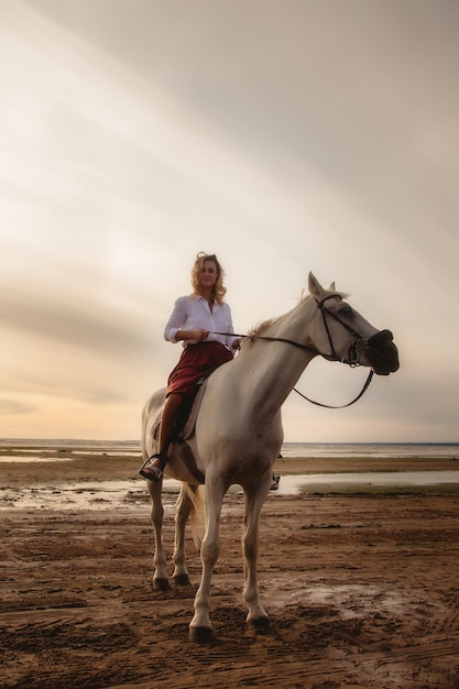 Ładny szczęśliwy młoda kobieta na koniu w lato plaża nad morzem. Kobieta jeździec jeździ swoim koniem w przyrodzie na tle zachodu słońca wieczorem światło. Koncepcja jazdy na świeżym powietrzu, sportu i rekreacji. Skopiuj miejsce