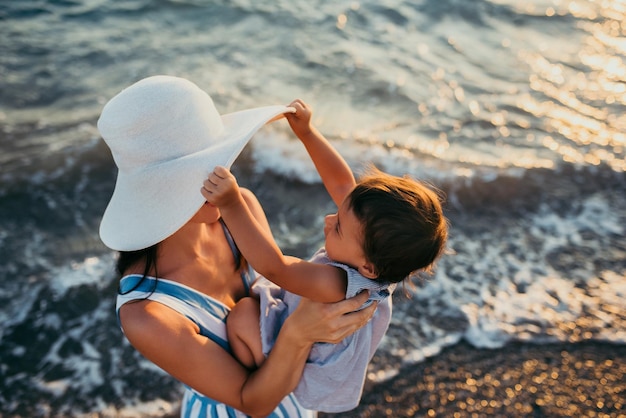 Ładny widok z góry młodej matki bawiącej się z córką w białym kapeluszu na tle morza Rodzinne wakacje Podróże Kobieta bawi się z wesołym maluchem Kobieta rasy kaukaskiej z dzieckiem o zachodzie słońca na oceanie
