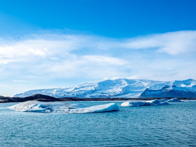 Laguna lodowcowa Jokulsarlon z lodowcem i dużą górą lodową pod zachmurzonym błękitnym niebem na Islandii