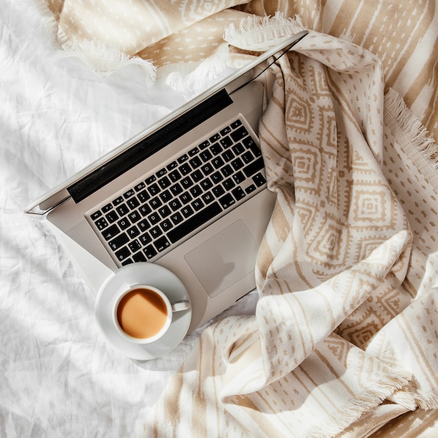 Laptop i filiżanka kawy na białym łóżku z beżową kratą. Praca w domu koncepcja. Poranne światło . Koncepcja stylu życia