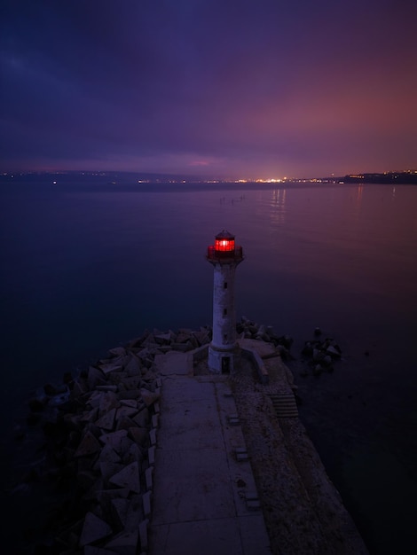 Latarnia morska na brzegu morza z czerwonym światłem w nocy na tle świateł statków
