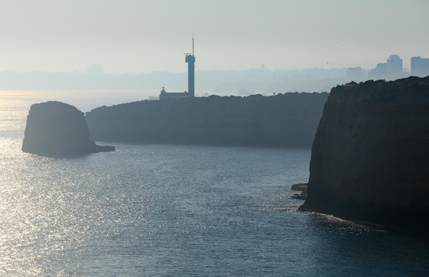 Latarnia morska Ponta do Altar na cyplu. Wieczorem wybrzeże atlantyckie mglisty widok z odbiciem słońca na powierzchni wody (Ferragudo, Lagoa, Algarve, Portugalia).