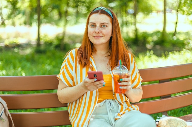 Lato piękna szczęśliwa kobieta ze smartfonem trzyma szklankę sody siedzącą na drewnianej ławce w mieście pa...