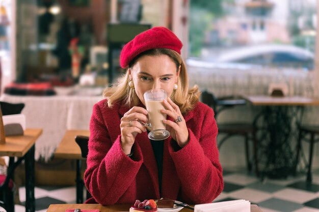 Zdjęcie latte w kafeterii. elegancka francuzka ubrana w ładne kolczyki i czerwony płaszcz pije latte w stołówce