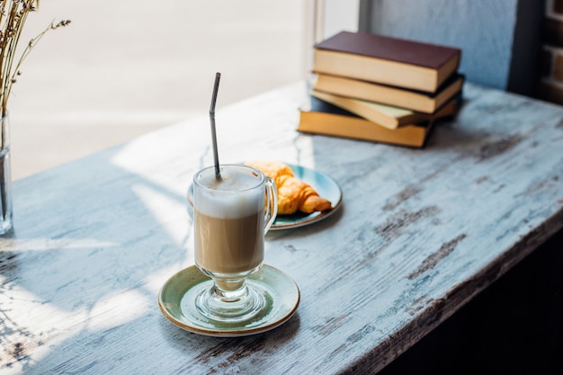 Latte w wysokiej szklance na tle stosu książek. Napój leży na stoliku w kawiarni, obok rogalika. Pyszne śniadanie i przekąska. Przyjemny wypoczynek i rekreacja.