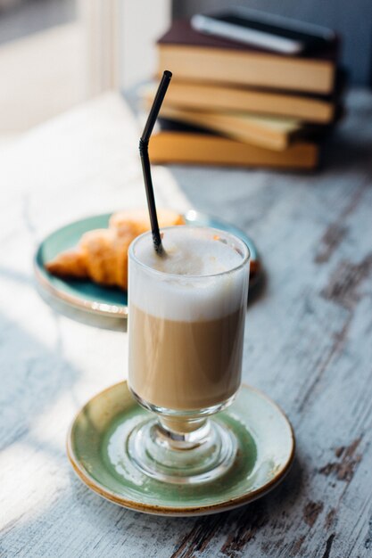 Latte w wysokiej szklance na tle stosu książek. Napój leży na stoliku w kawiarni, obok rogalika. Pyszne śniadanie i przekąska. Przyjemny wypoczynek i rekreacja.